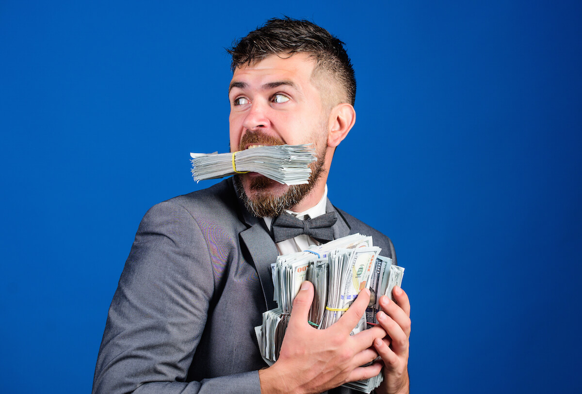 Man holding piles of money in his hands and mouth, looking over his shoulder as though someone will take it